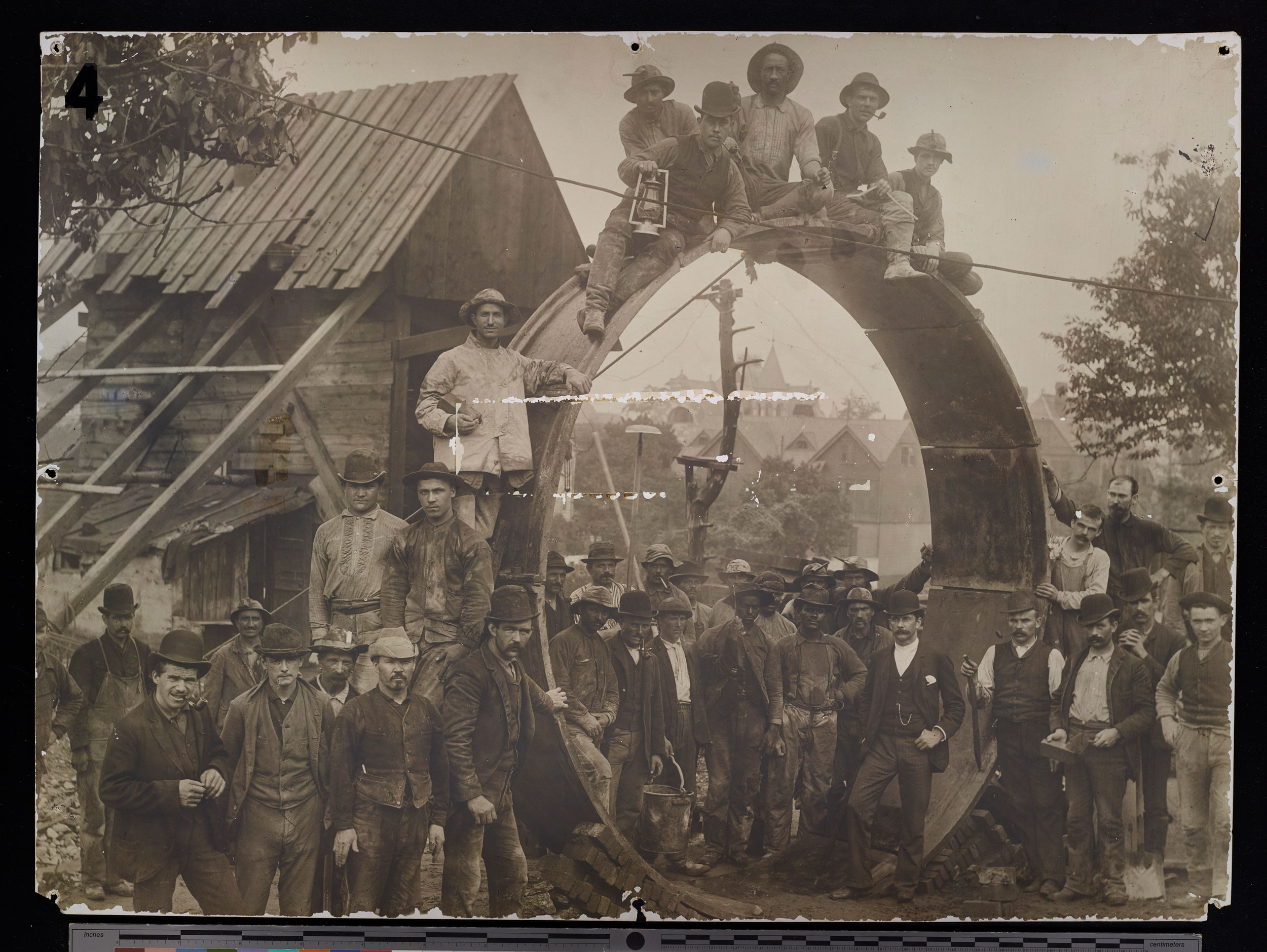 Photo showing workers in front of a wooden house and surrounding a circular gasket used for building the tunnel. Some workers are shown sitting on top of it and several others stand inside of it.