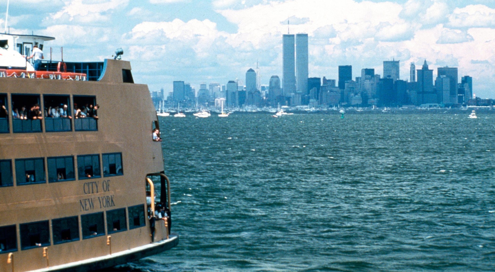 Color photograph of New York City waterfront showing a ferry boat in the left and a view of lower Manhattan.
