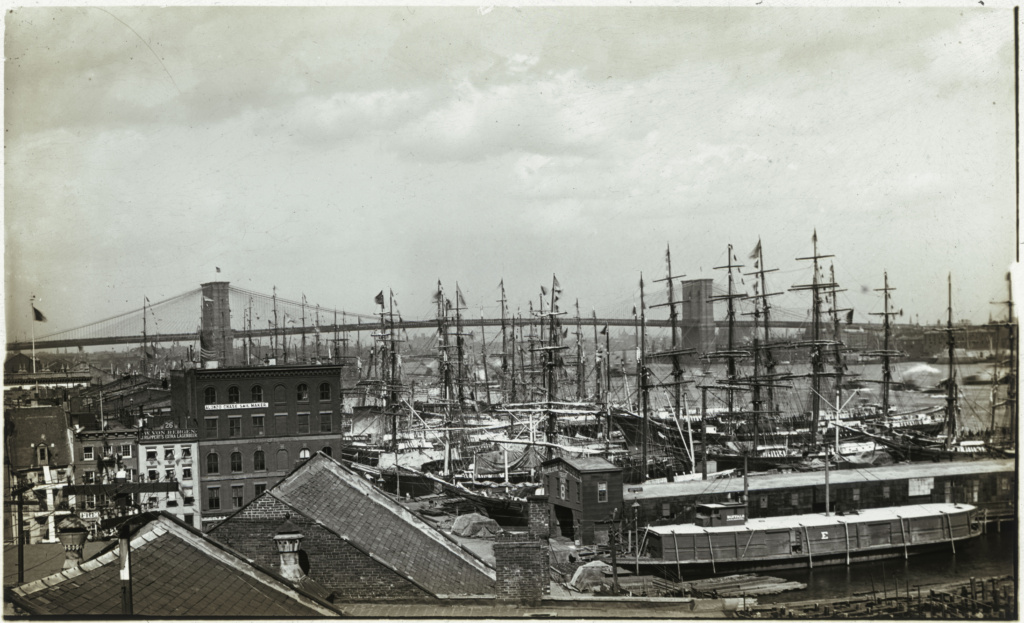 Photograph of a wharf with ships in port and the Brooklyn Bridge in background
