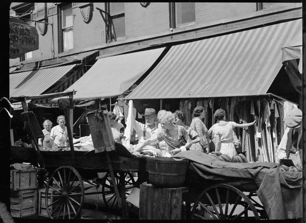 Black and white photograph displaying customers buying fruit and clothing at pushcarts parked on the curb. Behind the carts there are women looking at clothing hung in a store with a large awning.
