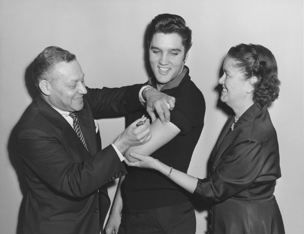 Black and white photograph showing Elvis Presley getting a vaccine shot from a staff member while Health Commissioner Baumgartner holds his arm.