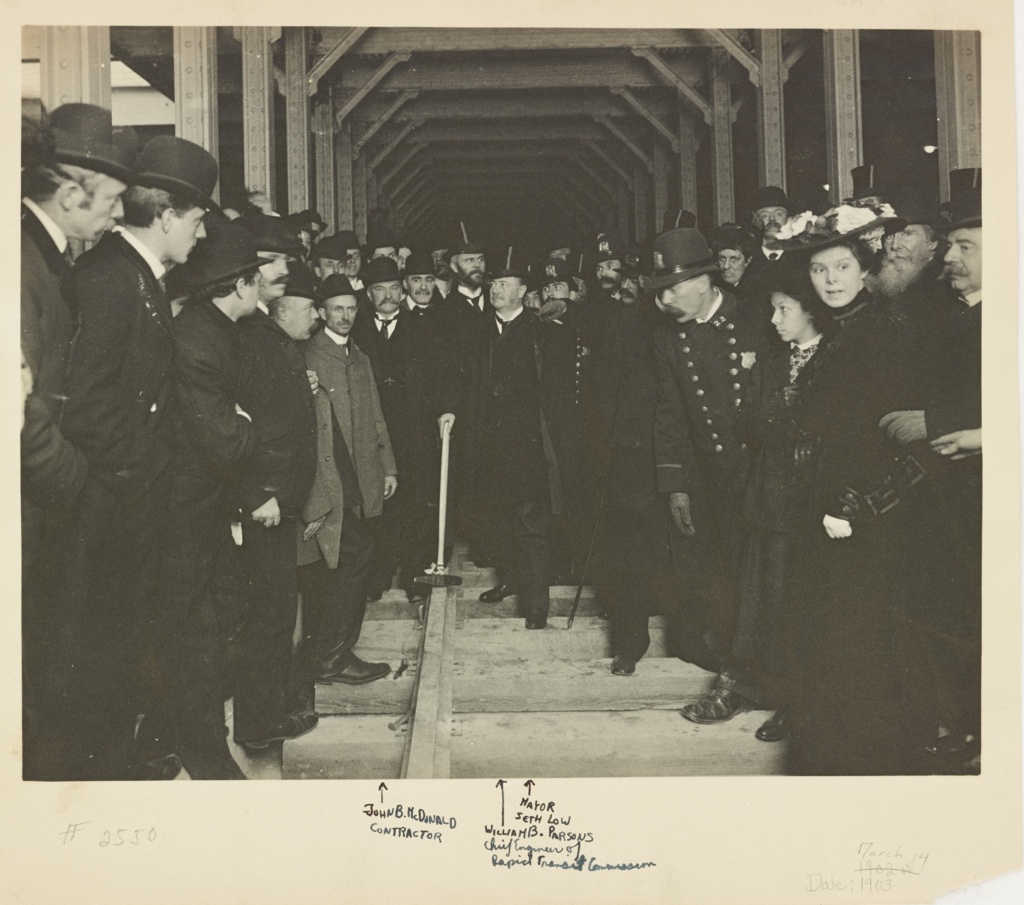 Group of men and women gathered at the entrance of a subway tunnel. One man holds a large hammer.The photo has inscriptions that identify John McDonald, Mayor Seth Low and William Parsons