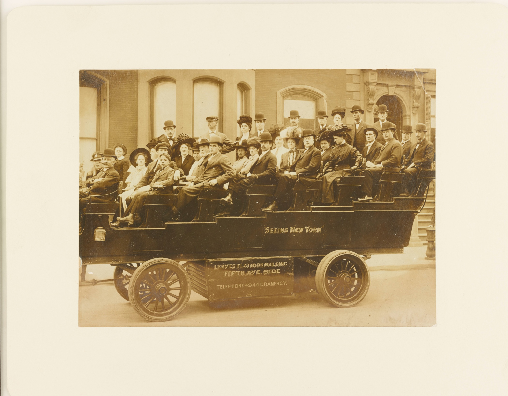 Sepia colored photograph shows people seated in a open air motorized bus with stadium seating. The sign on the bus reads "Seeing New York" and another sign reads, "Leaves Flatiron Building Fith Ave Side" and lists a telephone number.