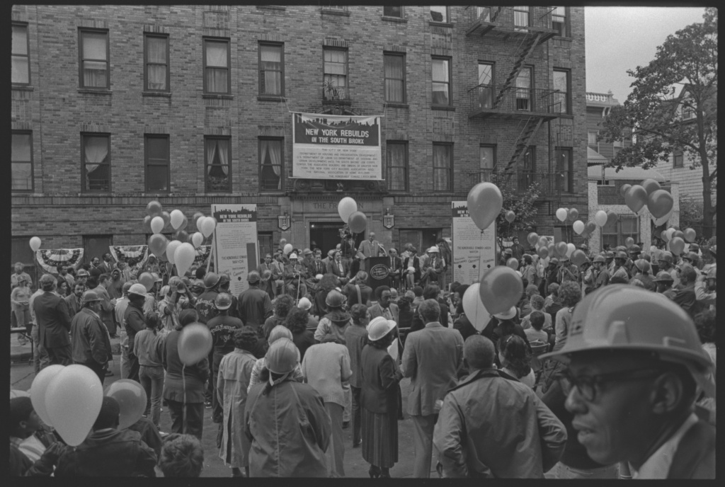 Black and white photo showing large crowd gathered and holding many balloons in front of building in the South Bronx.There is a stage in front of the building where someone is speaking.