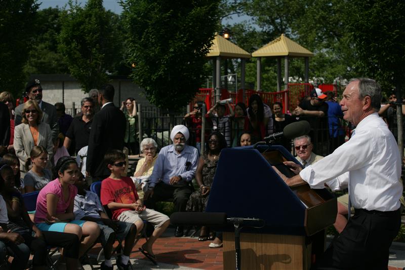 Color photograph of Mayor Bloomberg giving a speech at Elmhurst park with crowd of people.