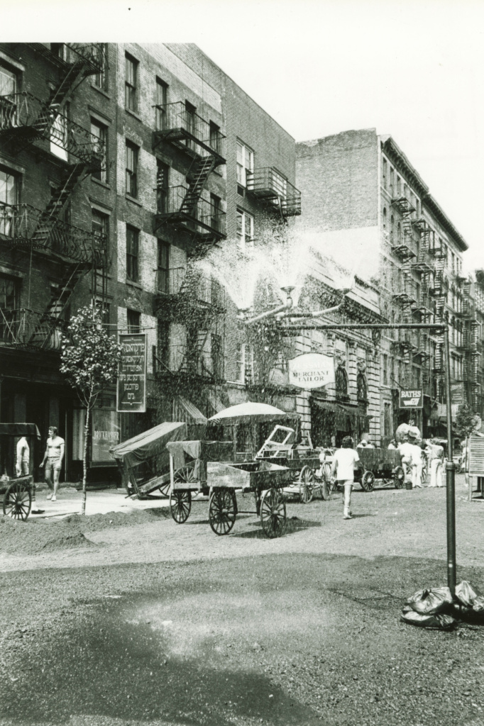 Black and white city scene showing some pushcarts and several businesses nearby including a tailor shop, one business with sign written in Hebrew and a bathouse facility. A tall multiheaded sprinkler stands in the street shooting up water.