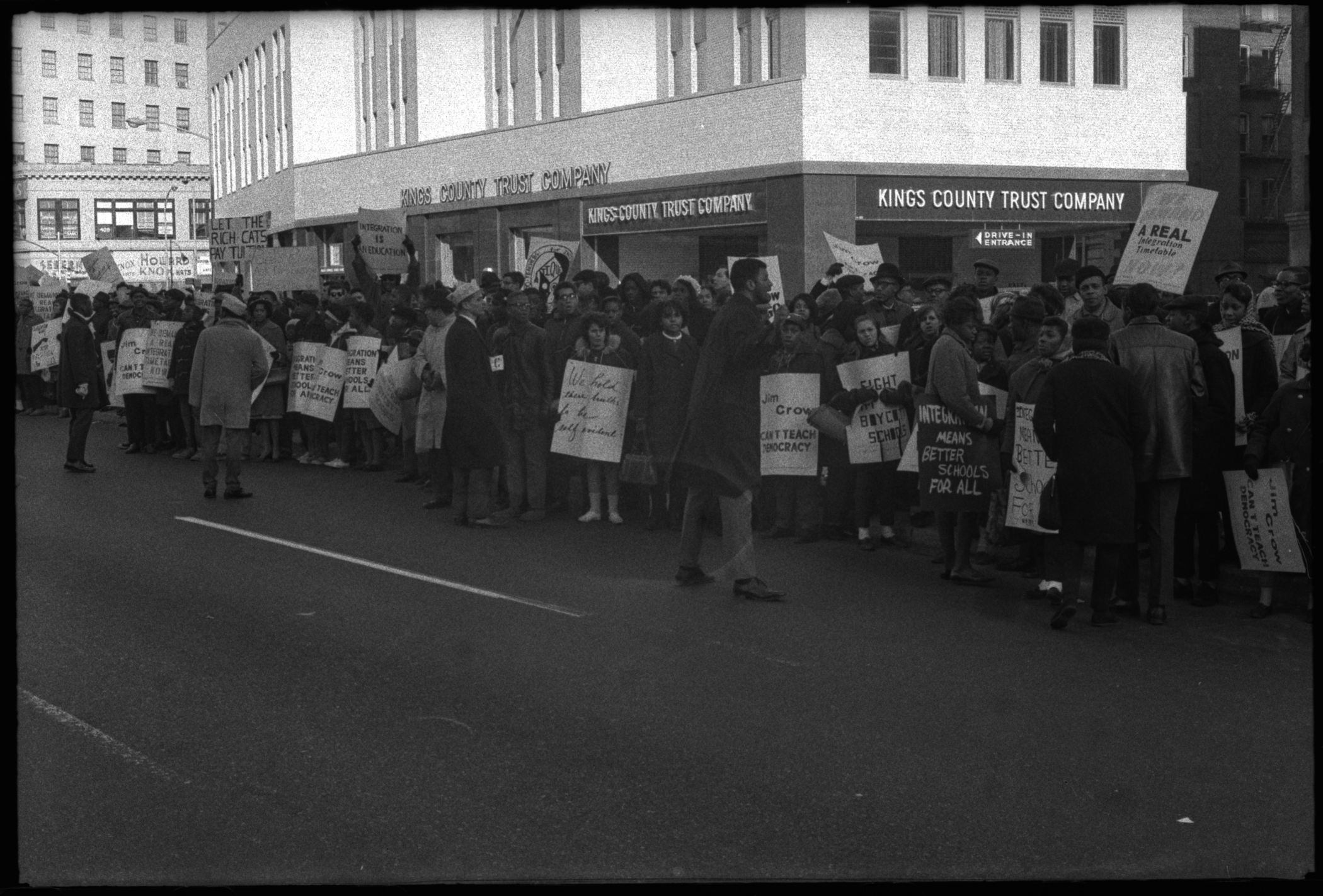 Photograph of a large crowd at a demonstration for school integration. The crowd includes students holding signs in front of the Kings County Trust Company.