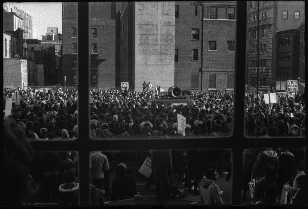 Black and white photograph of demonstrators crowding city street while a car with several loudspeakers is parked in the center.