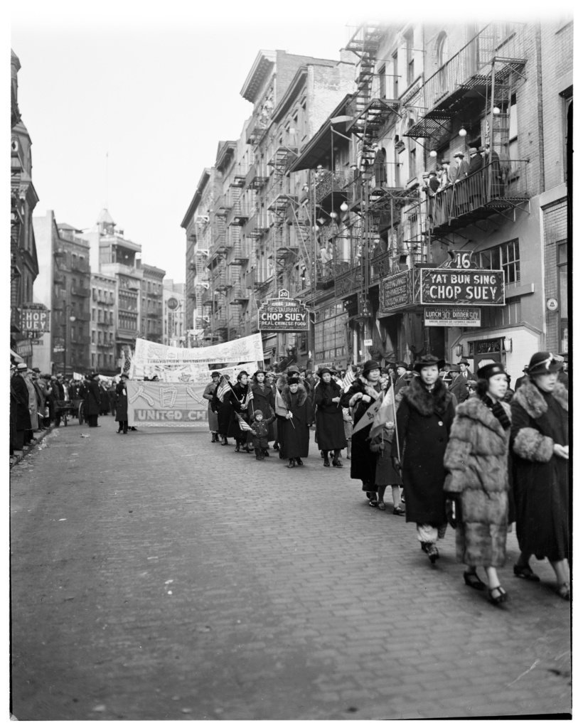 Photograph showing people, (mostly women) walking in parade carrying banners and Chinese and American flags down Mott Street.