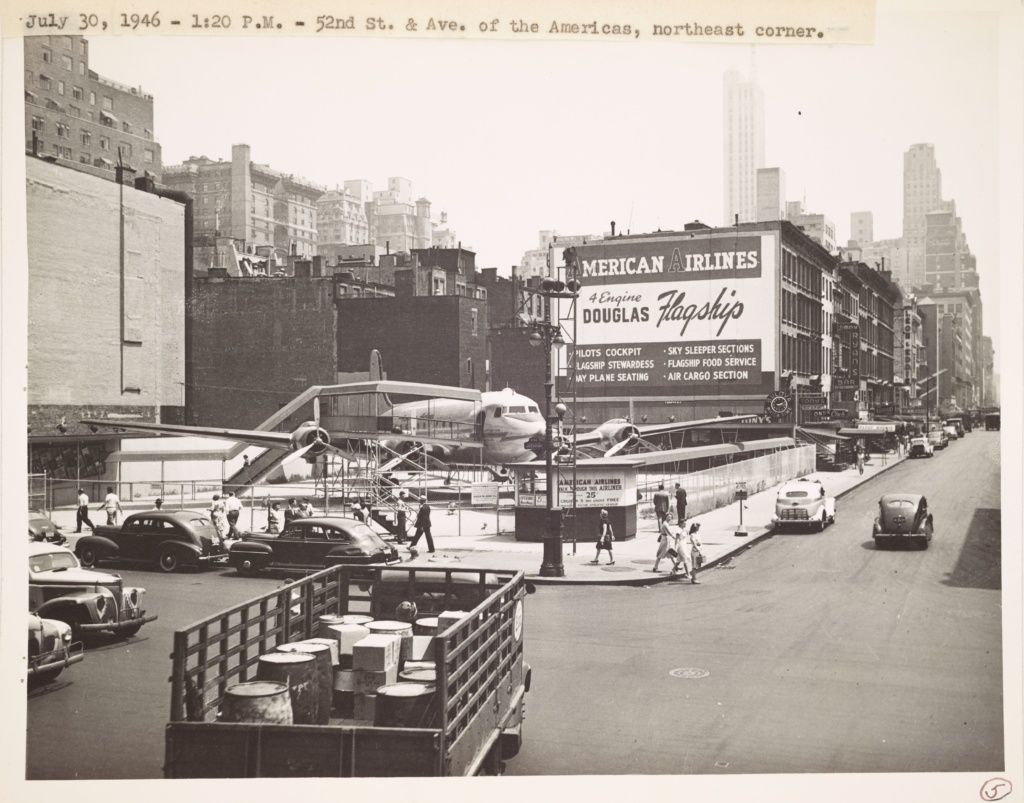 Photograph showing an airplane parked on corner of 52nd Street and 6th Avenue, surrounded by fencing. A billboard promotes the American Airline flagship. There are pedestrians on the sidewalk and crossing the street and parked cars.