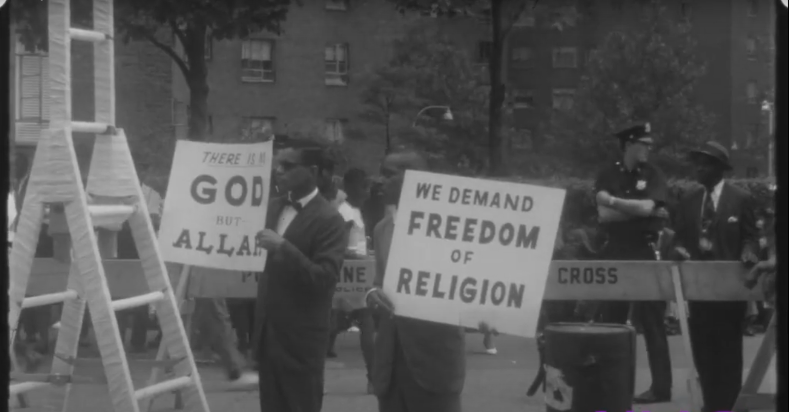 Screenshot from a video showing Malcolm X standing at a podium during an outdoor rally.