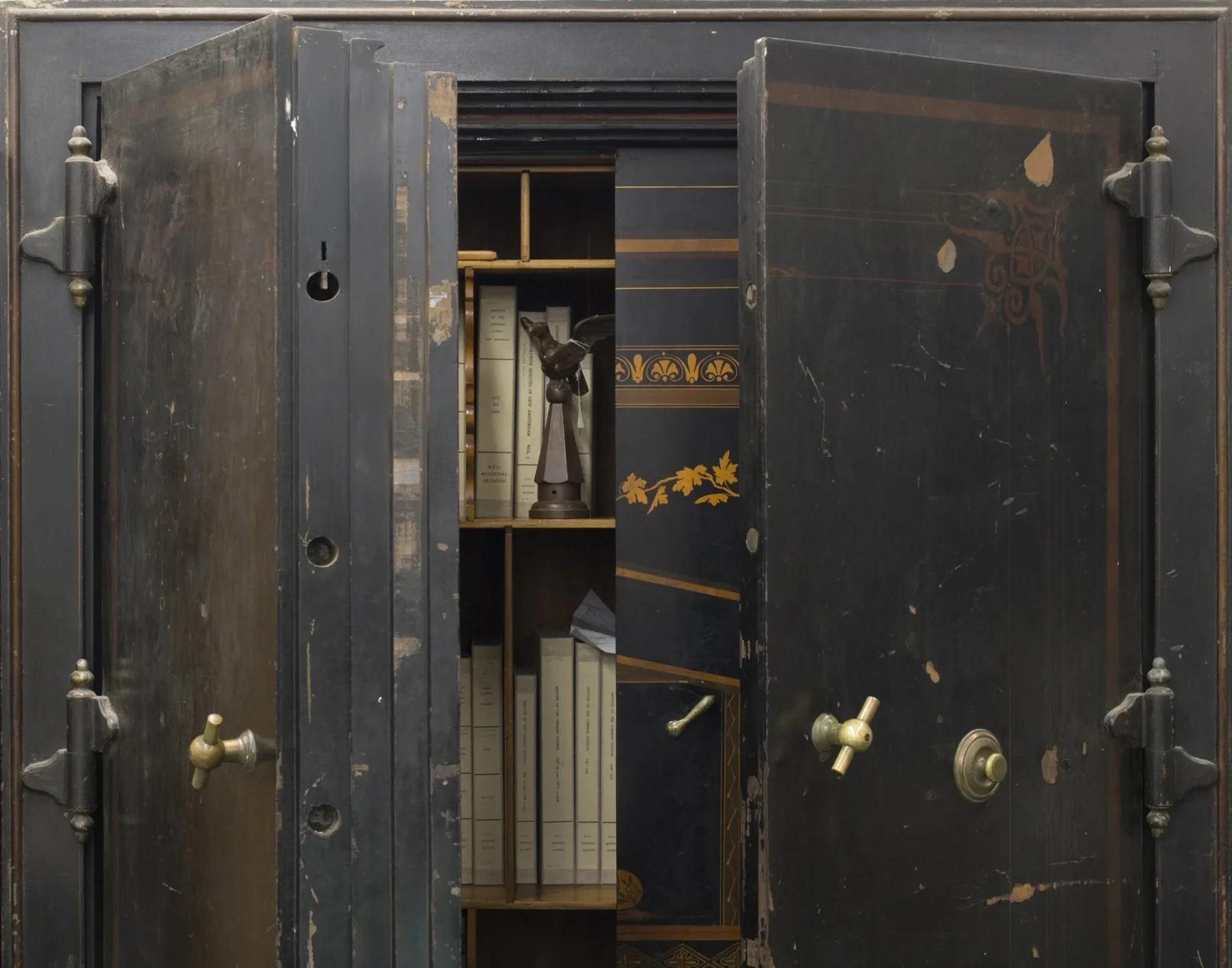 Large black safe with ornate painting partially open, showing interior shelves holding books, narrow boxes and a bird statuette. .