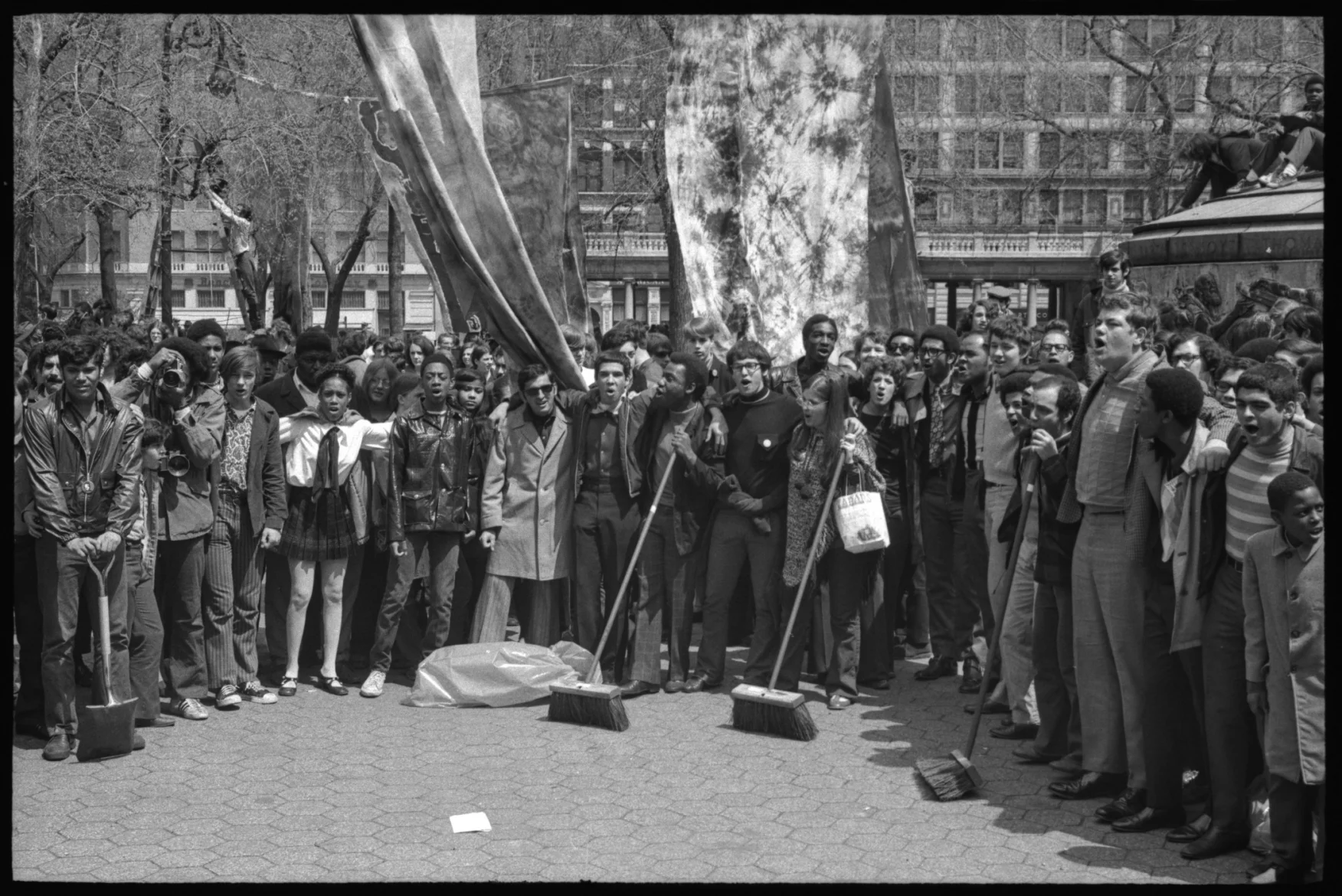 Black and white photograph displaying crowd at Union Square park. The crowd features people from different races and ages, some holding brooms.